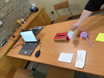An escape-room workstation set up with laptops, a clue dropbox, printed clue hints, scratch paper, and pens.